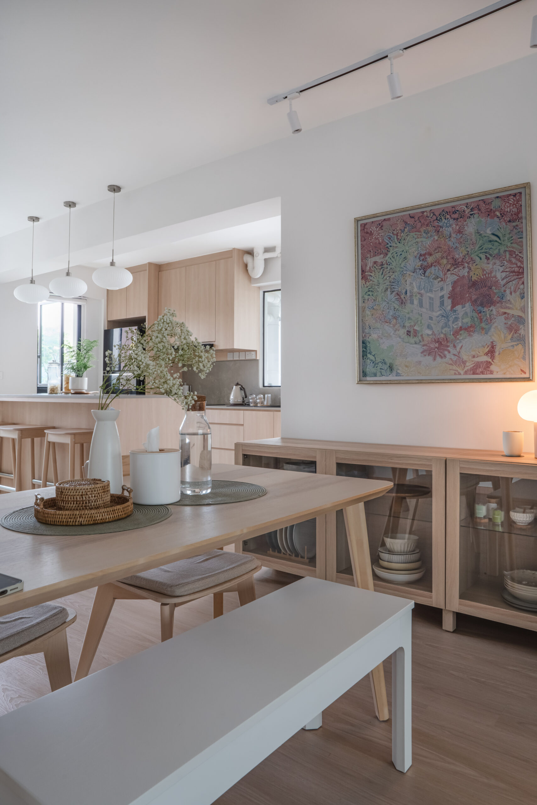 A stunning Singaporean interior design home featuring a modern Scandinavian-inspired kitchen with a beautiful blend of wood and white elements. The kitchen showcases sleek wooden cabinets, a pristine white countertop, and stylish pendant lights. Additionally, the dining table is strategically placed at the center of the living room, creating a harmonious and open layout. The light-filled space exudes a cozy and inviting ambiance, combining minimalist aesthetics with warm, natural textures for a truly captivating design.